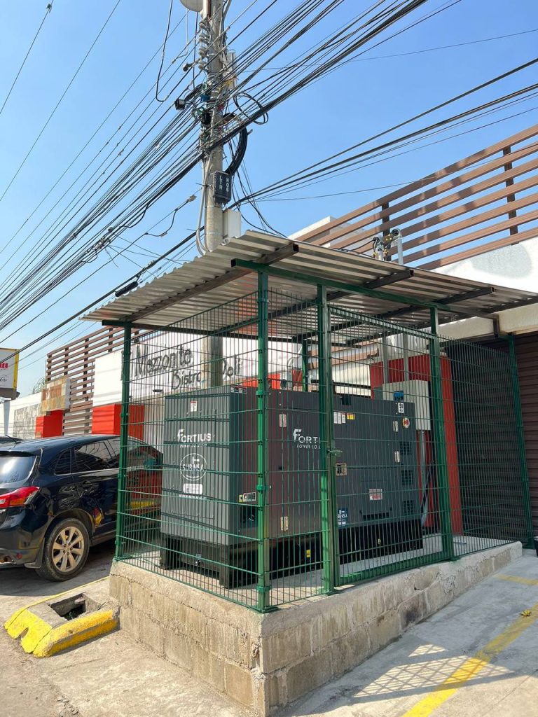 Generador eléctrico Fortius dentro de una estructura protectora hecha a base de varandales de hierro y un techo de lámina. Equipo se encuentra al frente de un restaurante llamado Mezzonite Deli & Bistro.